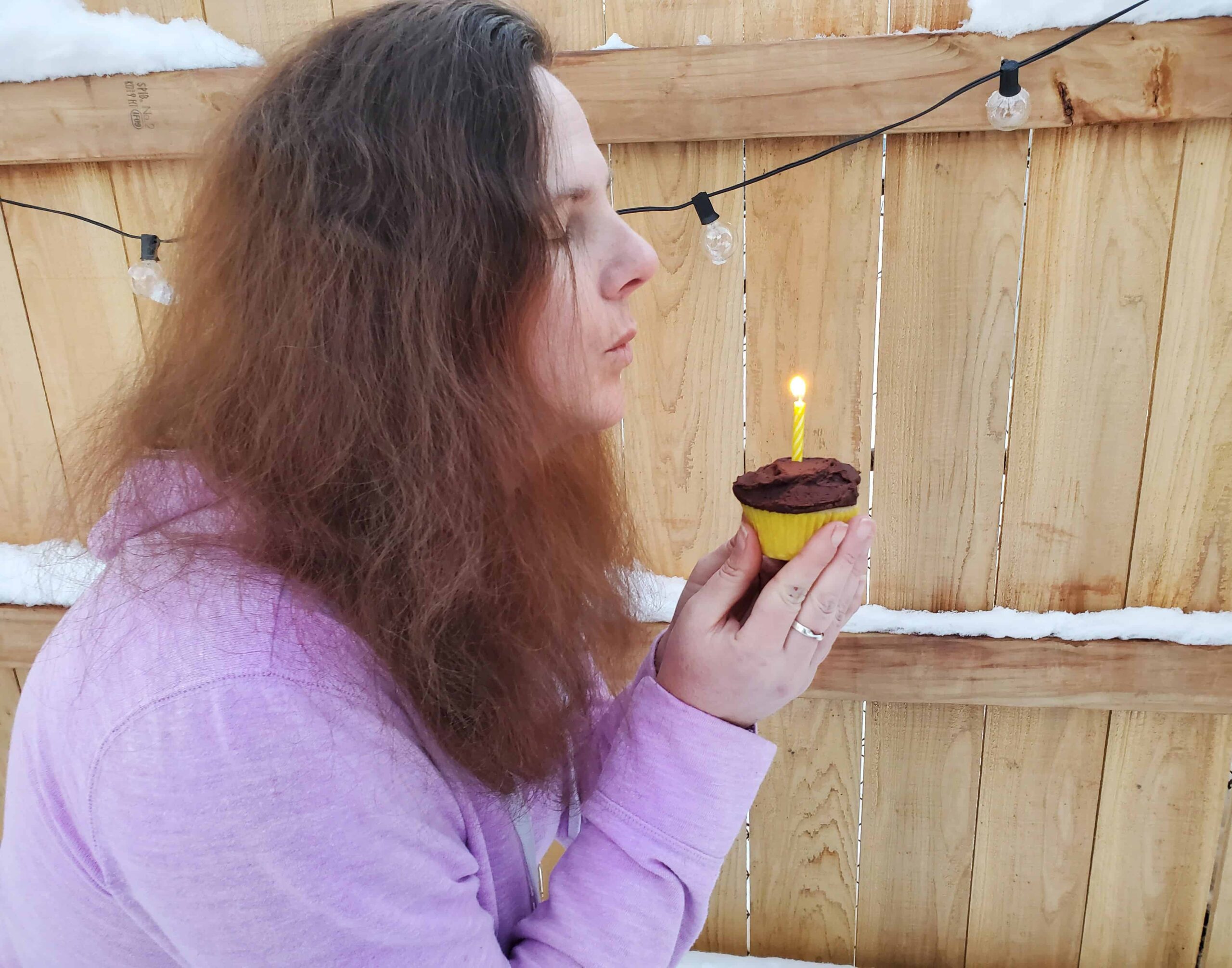 Woman blowing out a candle on a chocolate frosted cupcake by a wooden fence.