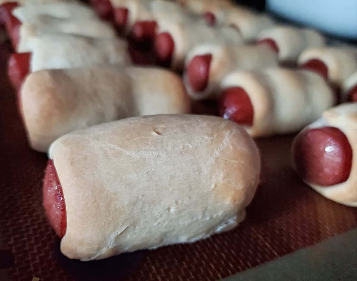 Image shows Hot dogs wrapped in crescent roll dough on a baking sheet.