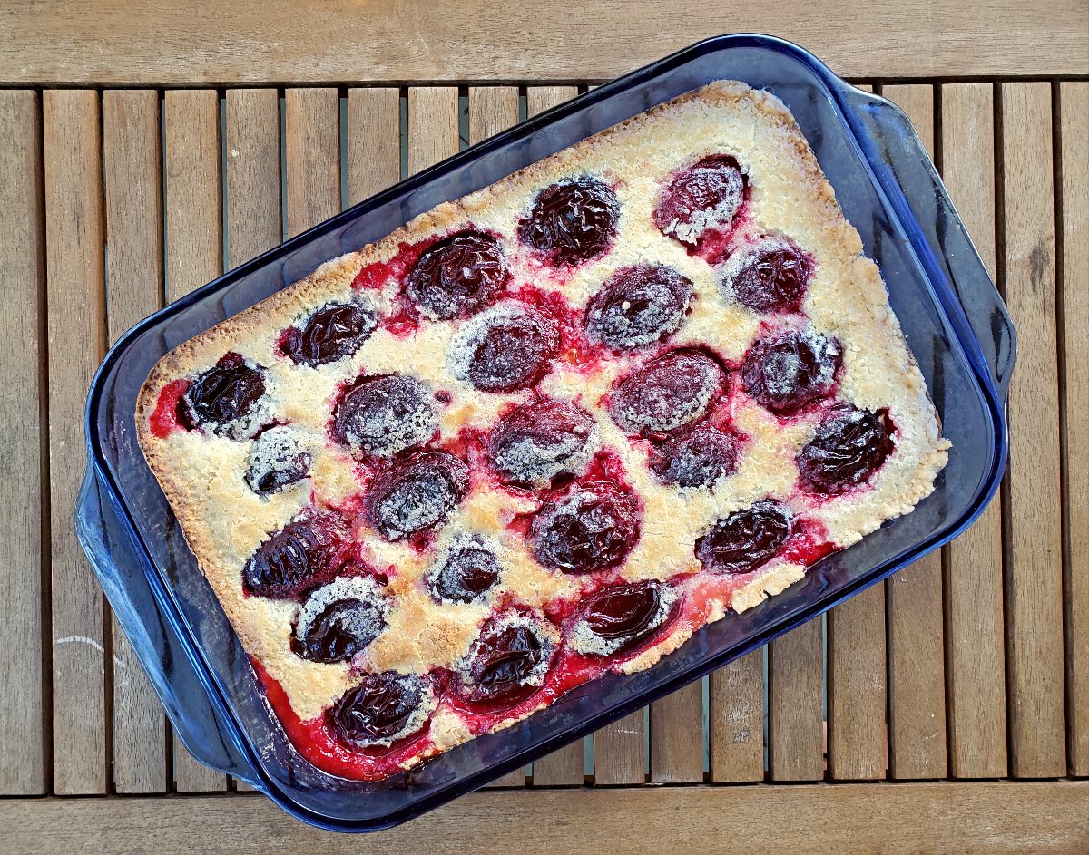 Image shows an overhead shot of German Plum Kuchen in a blue pyrex pan on a wooden table.