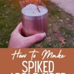 Hand holding whipped cream topped drink in front of a fire with text how to make spiked apple cider.