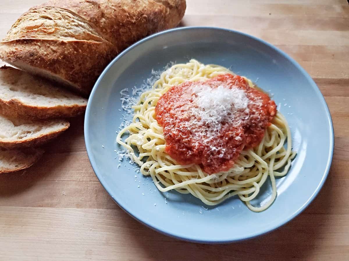 Blue plate of fresh pasta with sauce and cheese with a sliced loaf of bread to the left.