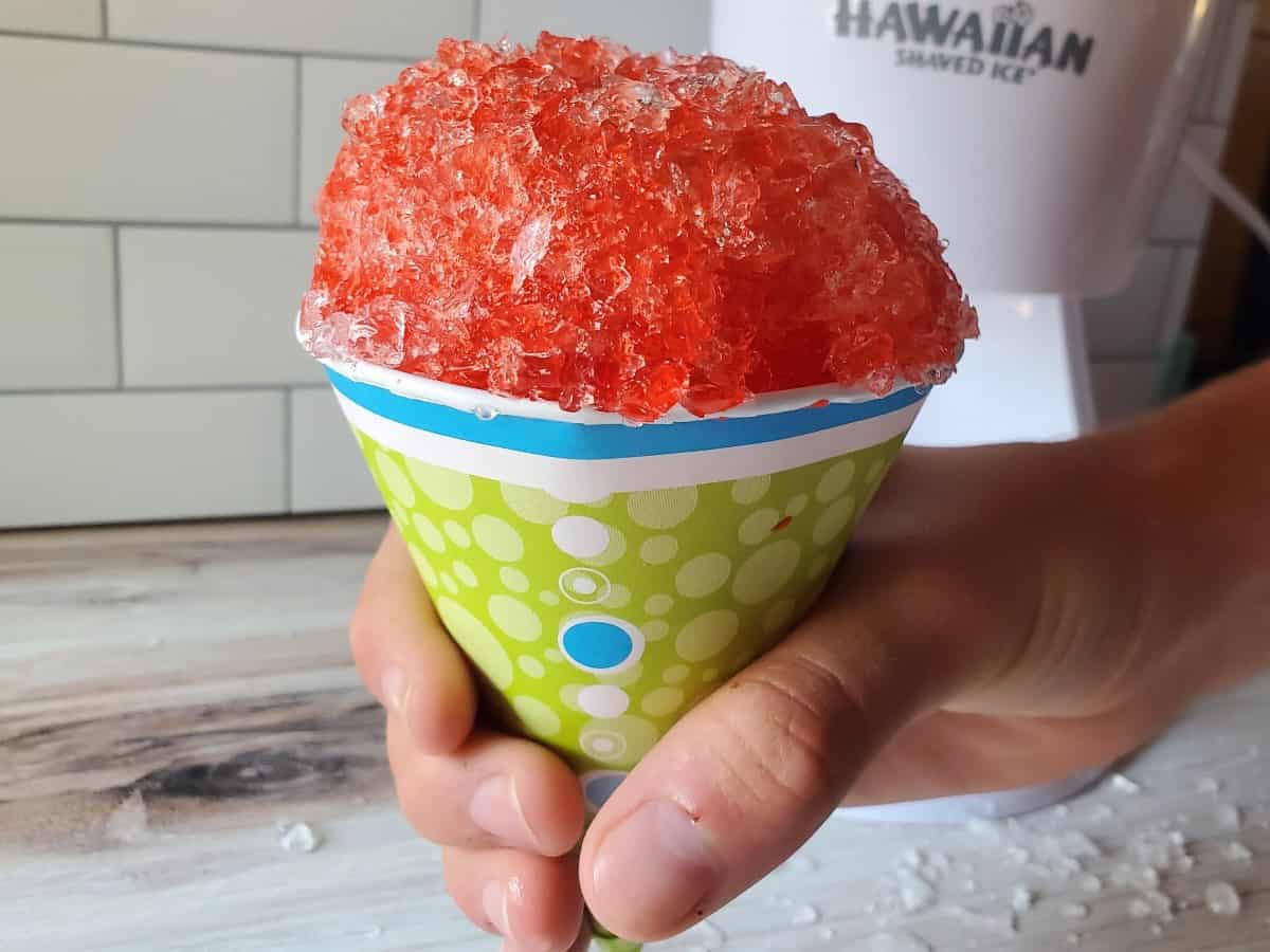 Hand holding a red snow cone in front of a shaved ice machine.