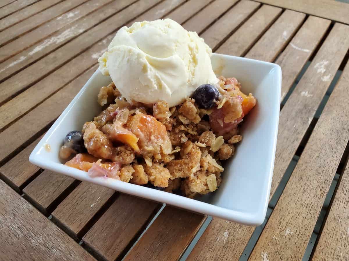 Image shows a white bowl with a serving of blueberry peach crisp topped with a scoop of ice cream and some whipped cream.