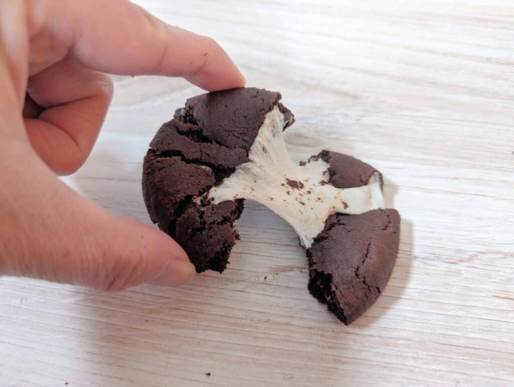 Hand pulling up half a gooey chocolate marshmallow cookie while it rests on a wooden board.