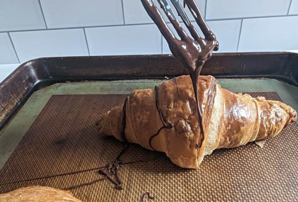 Fork drizzling chocolate onto a croissant on a baking tray.