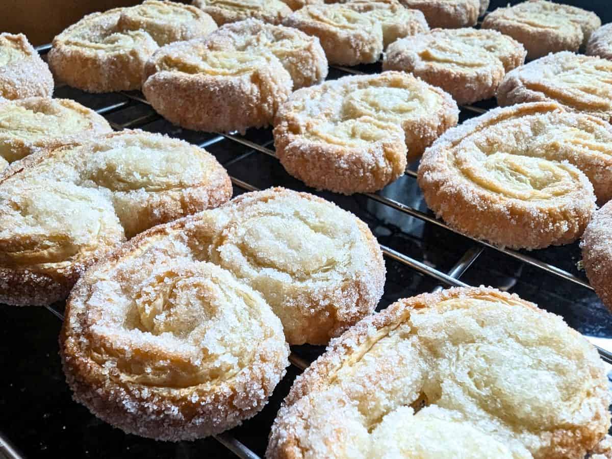 Image shows a hand holding a palmier cookie in front of others on a wire cooling rack.