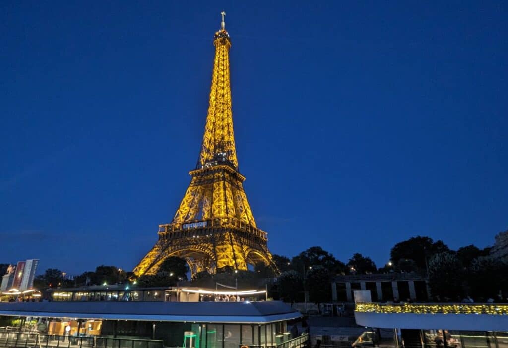 Image shows Eiffel Tower lit up at night from the vantage of the Seine River.