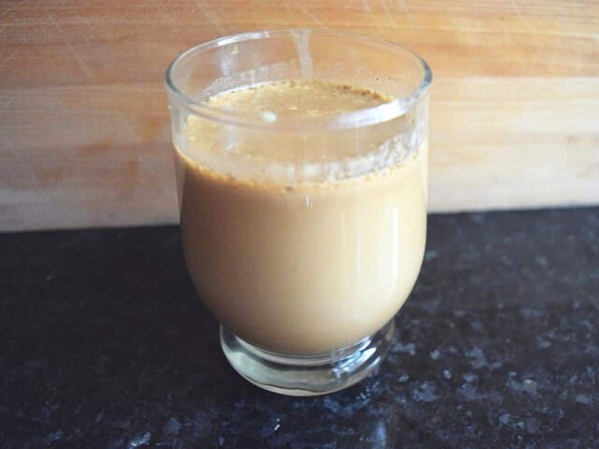 Image shows A glass and a bottle filled with a pumpkin spice latte are placed on a dark surface against a wooden backdrop.
