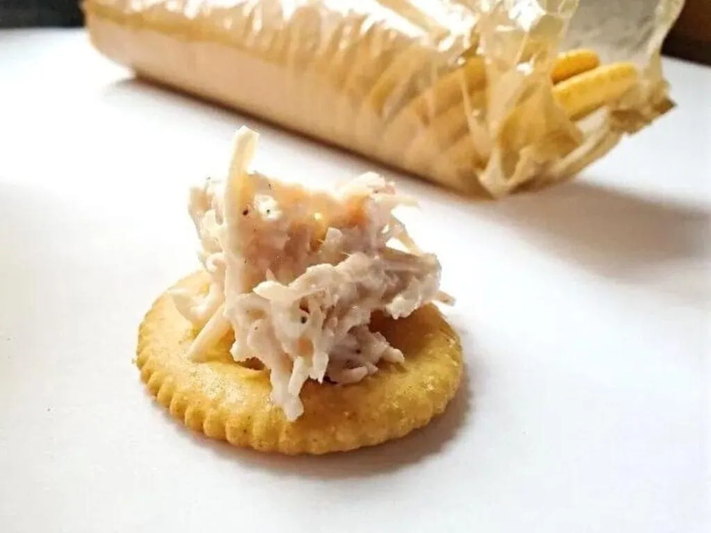 Image shows canned chicken salad on a cracker with more crackers in a package behind it.