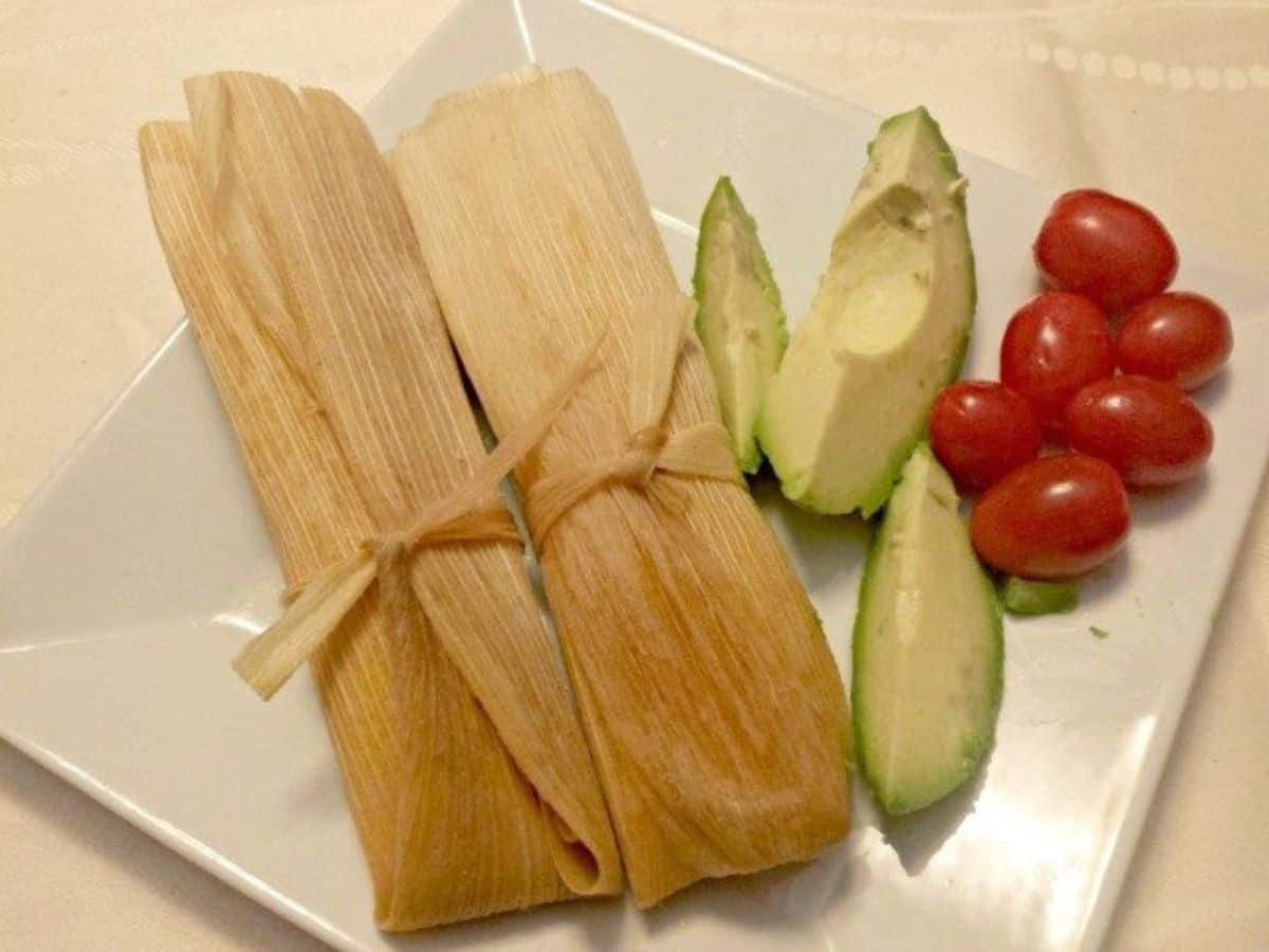 Image shows two Tamales sitting on a white plate with slices of avocado and tomatoes next to it.