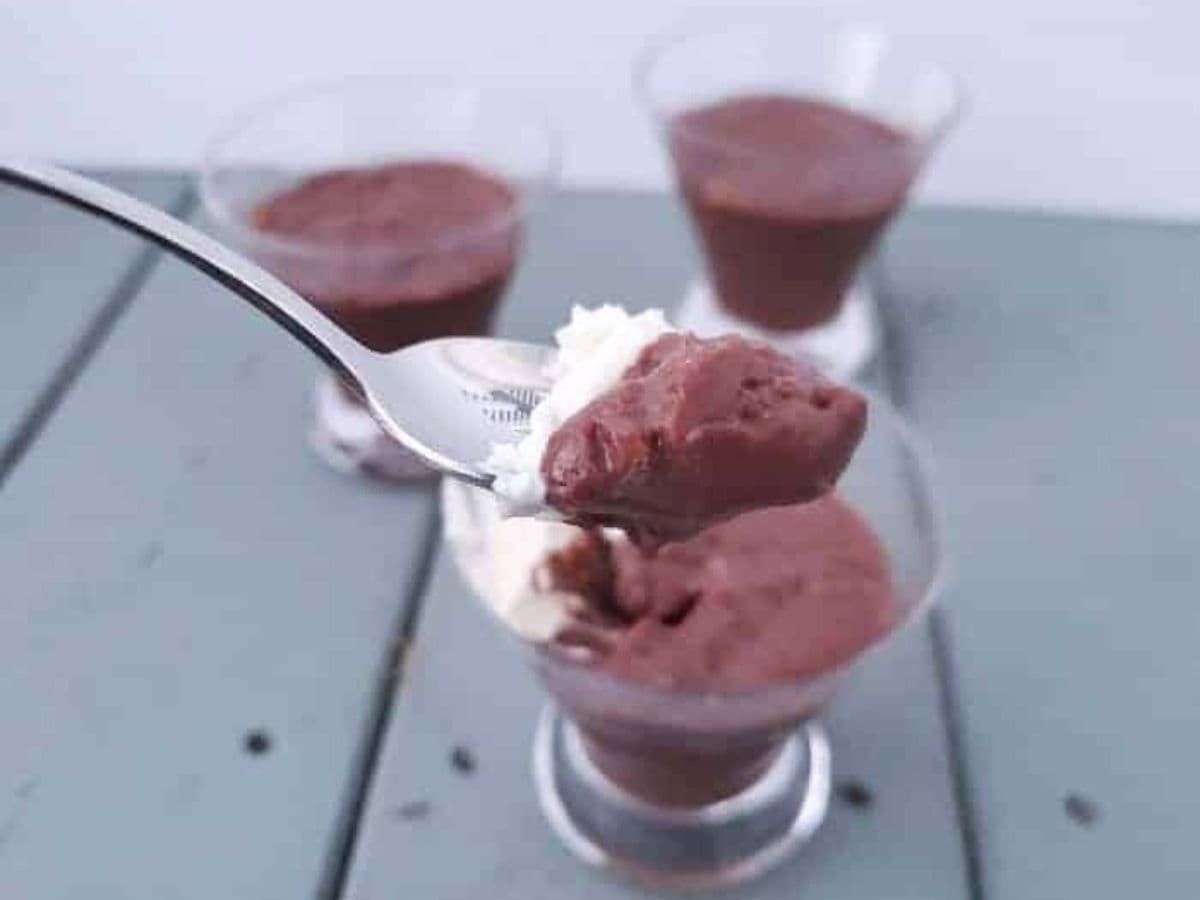 Image shows A close-up of a spoon with chocolate pudding above a glass filled with the same pudding. Three more glasses of pudding are in the background on a gray surface.