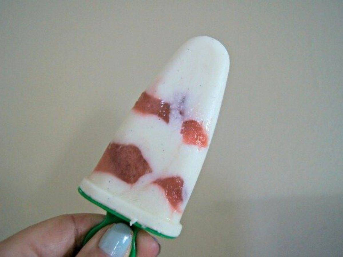 Image shows a hand holding a Greek yogurt popsicle against a plain background.
