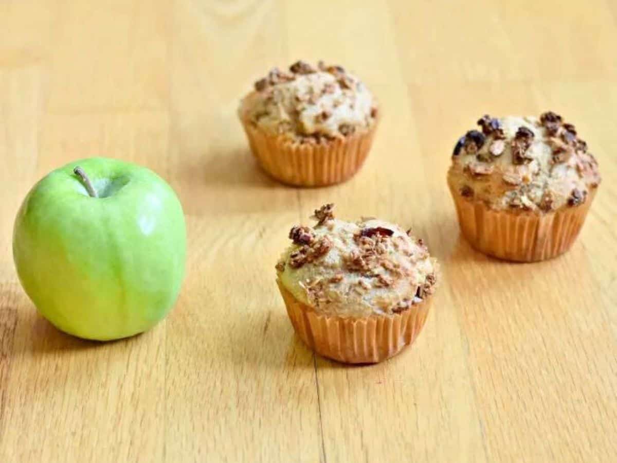 Three apple cinnamon muffins on a wooden table with an apple.