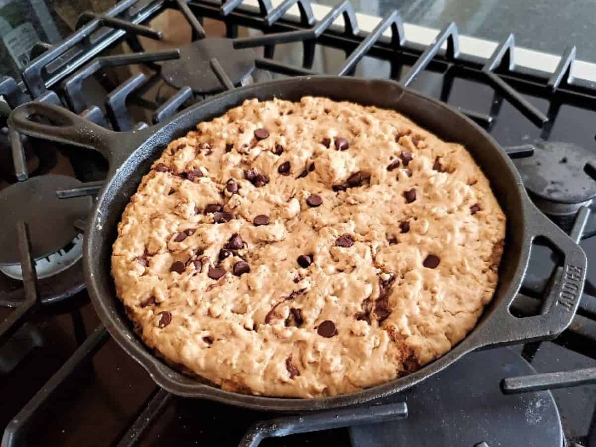 Image shows A piece of chocolate chip skillet cookie in front of a cast iron pan.