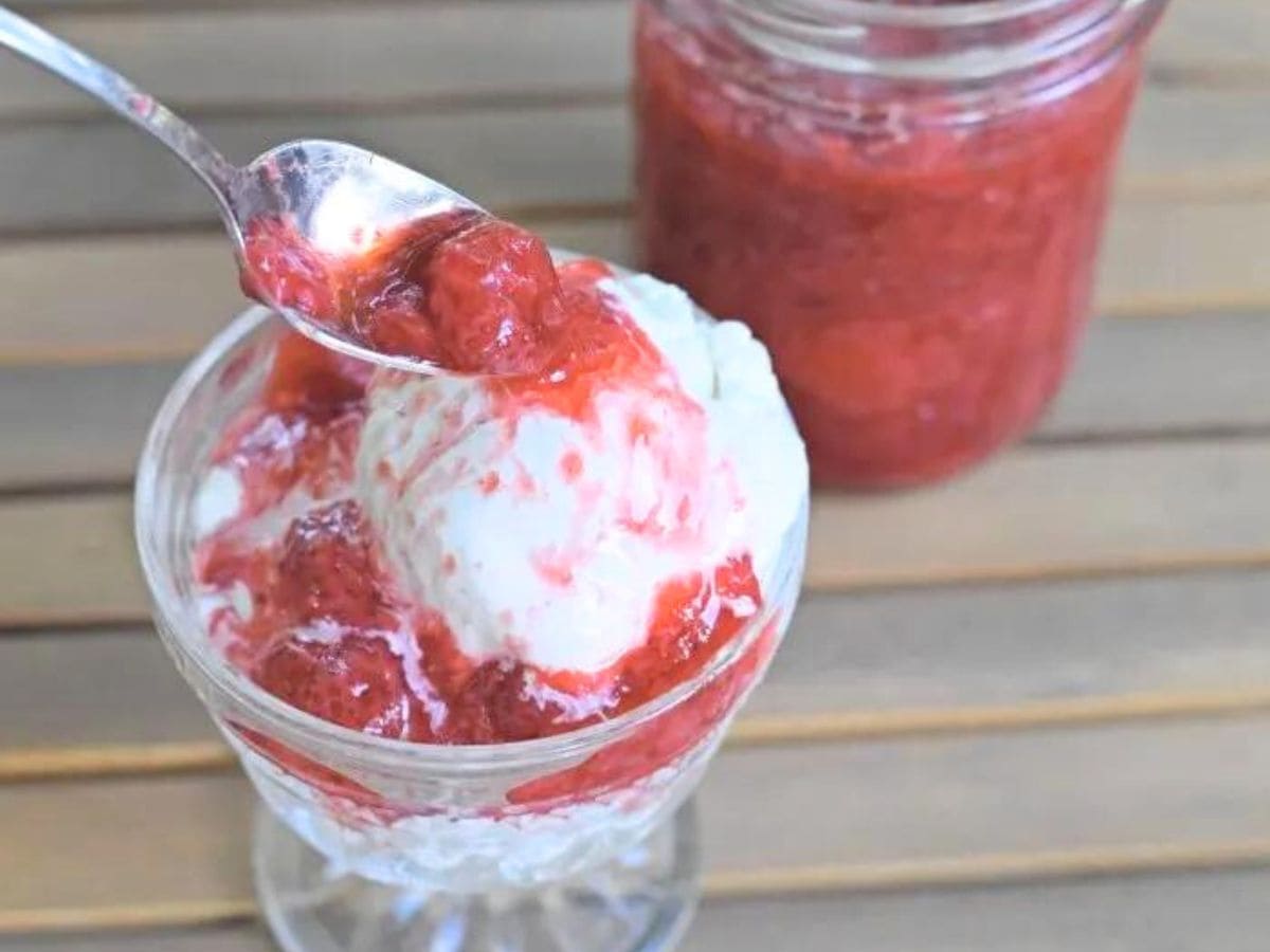 Spoon adding strawberry syrup to a glass of ice cream with a jar of strawberry syrup behind it.