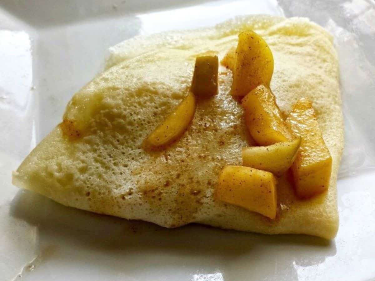 Image shows a close up of Cinnamon Apple Crepes with apples on top of a crepe folded into quarters.