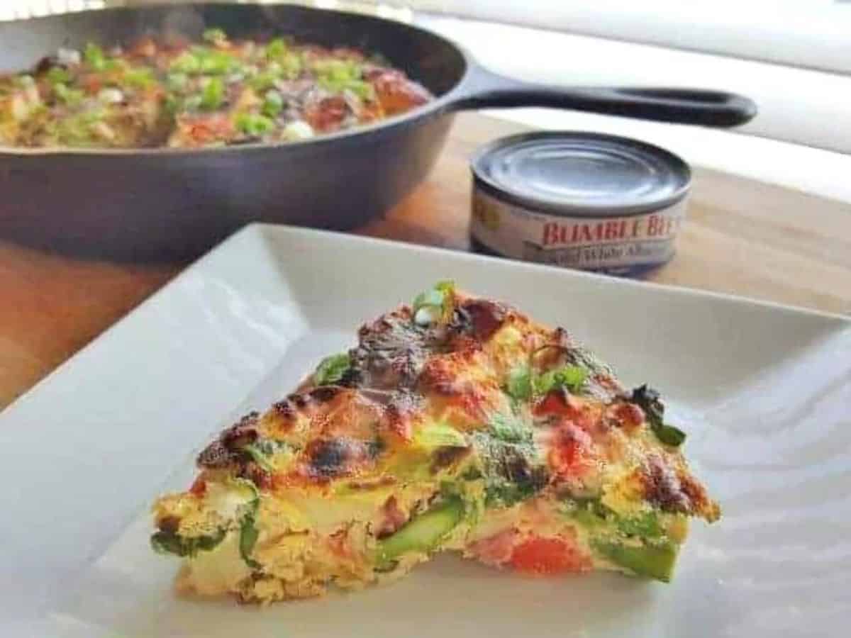 Image shows a slice of a Nicoise Omelet on a white plate with the cast iron skillet behind it.