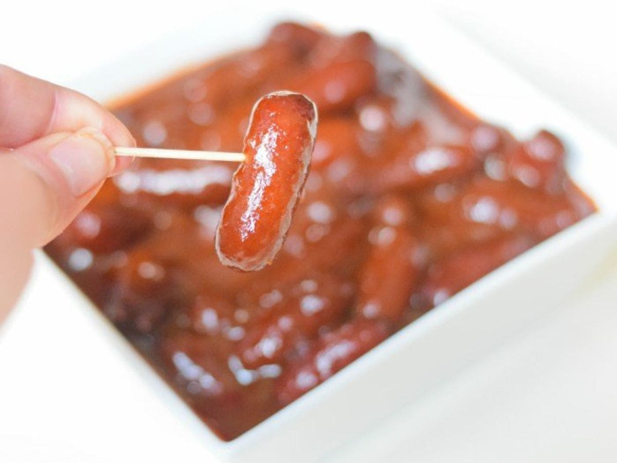 Image shows A person is holding a toothpick with a little smokie over a bowl filled with more bbq bourbon little smokies.