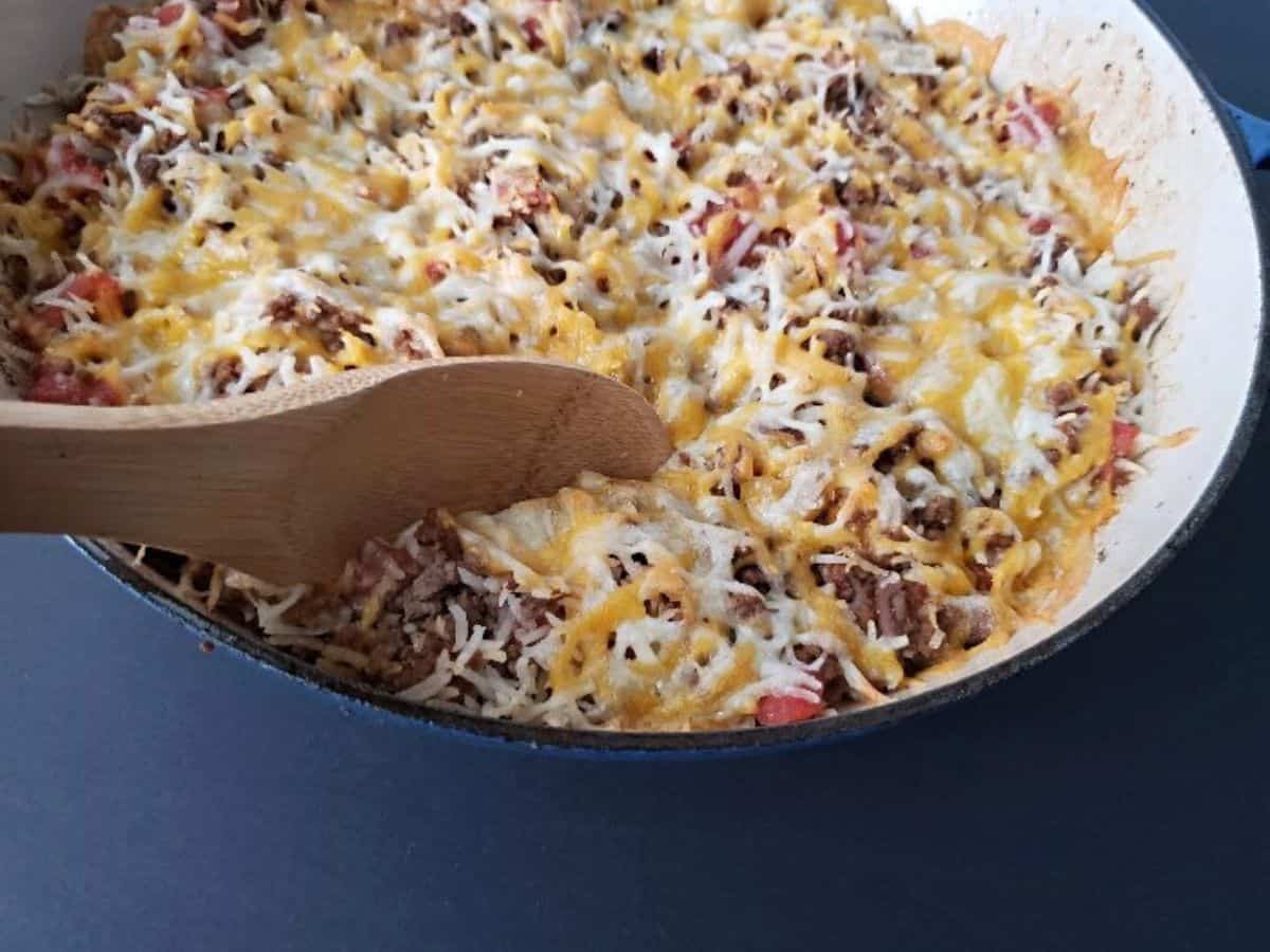 Image shows a closeup of a spoon holding some Cheesy Beef and Rice Casserole with the full skillet behind it.