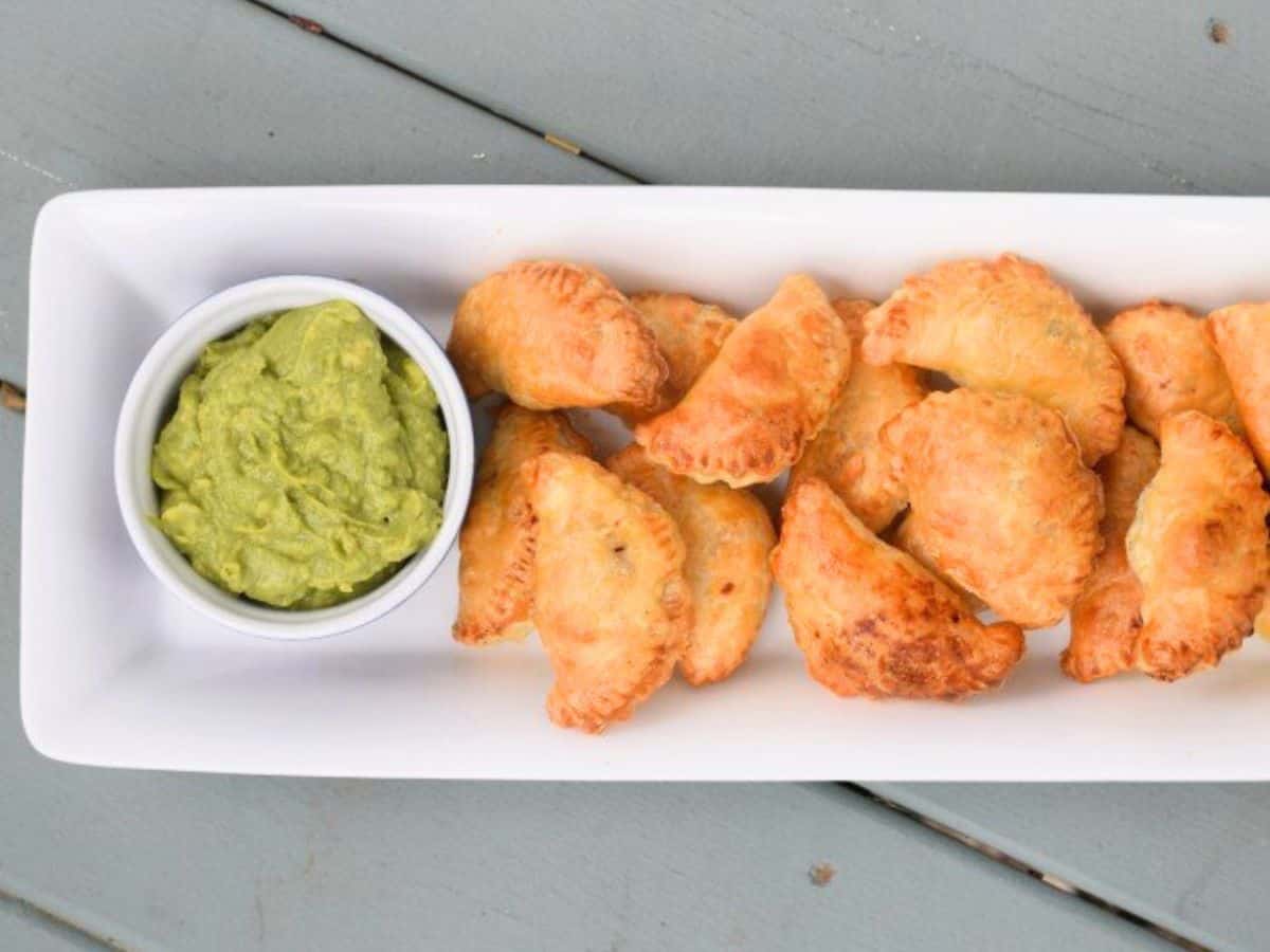 Image shows an overhead shot of Puff Pastry Beef Empanadas on a white tray with a bowl of guacamole to the left of them.
