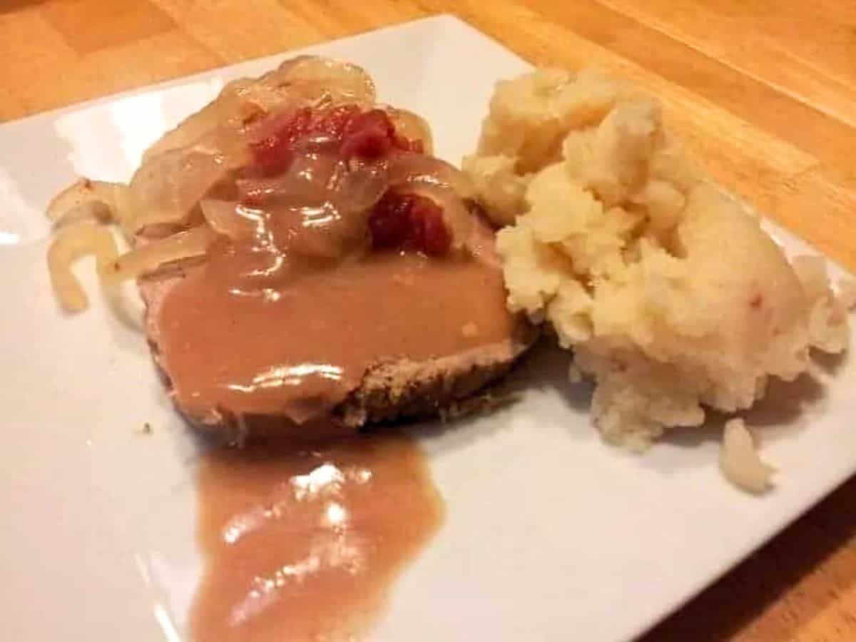 A plate of sliced roast meat topped with gravy, sautéed onions, and a few diced tomatoes, accompanied by a serving of mashed potatoes.