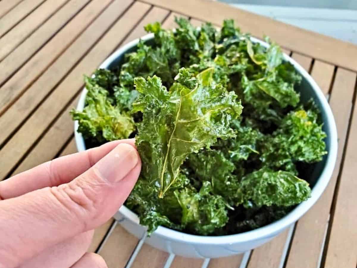 Image shows a hand holding a baked kale chip over a bowl filled with more kale chips.