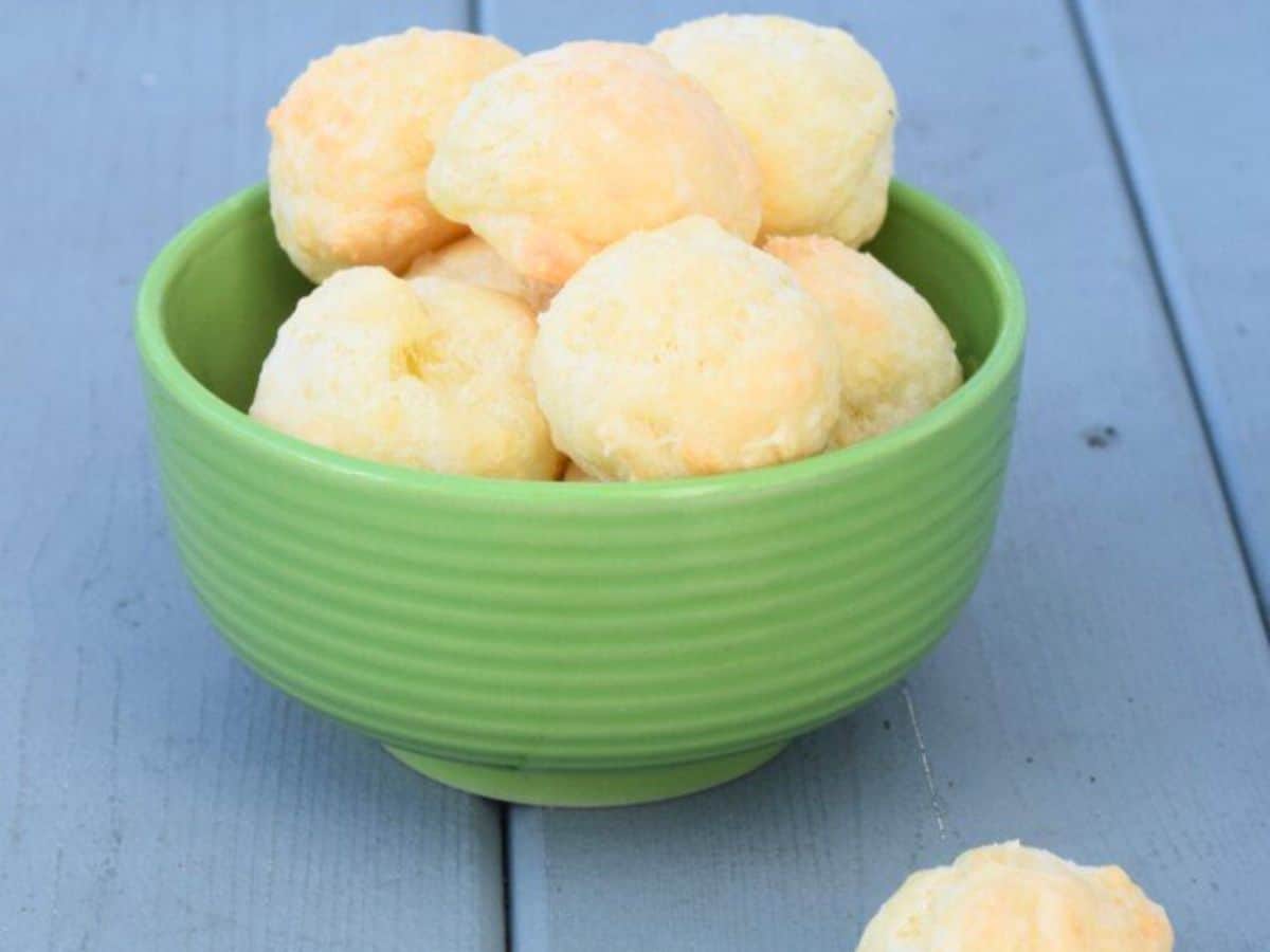 Hand breaking apart brazilian cheese bread with more in a bowl behind it.