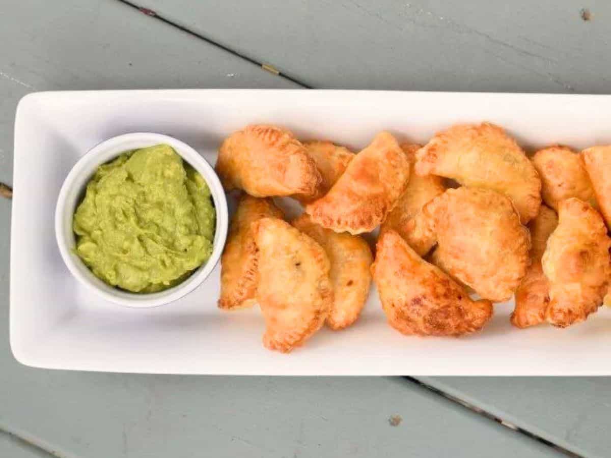 Image shows an overhead shot of Puff Pastry Beef Empanadas on a white tray with a bowl of guacamole to the left of them.