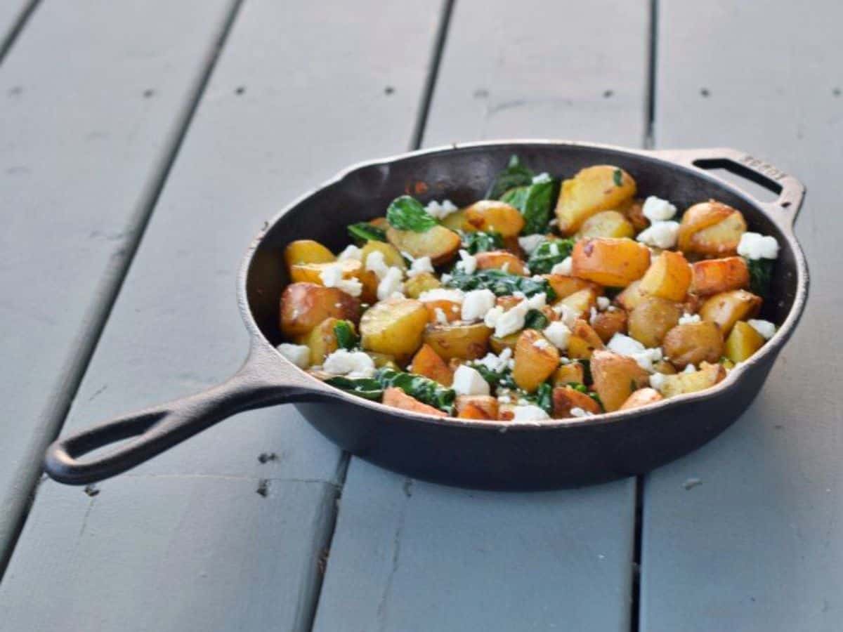 This photo shows a cast iron skillet with oven roasted potatoes with spinach and garlic on a blue wooden background.