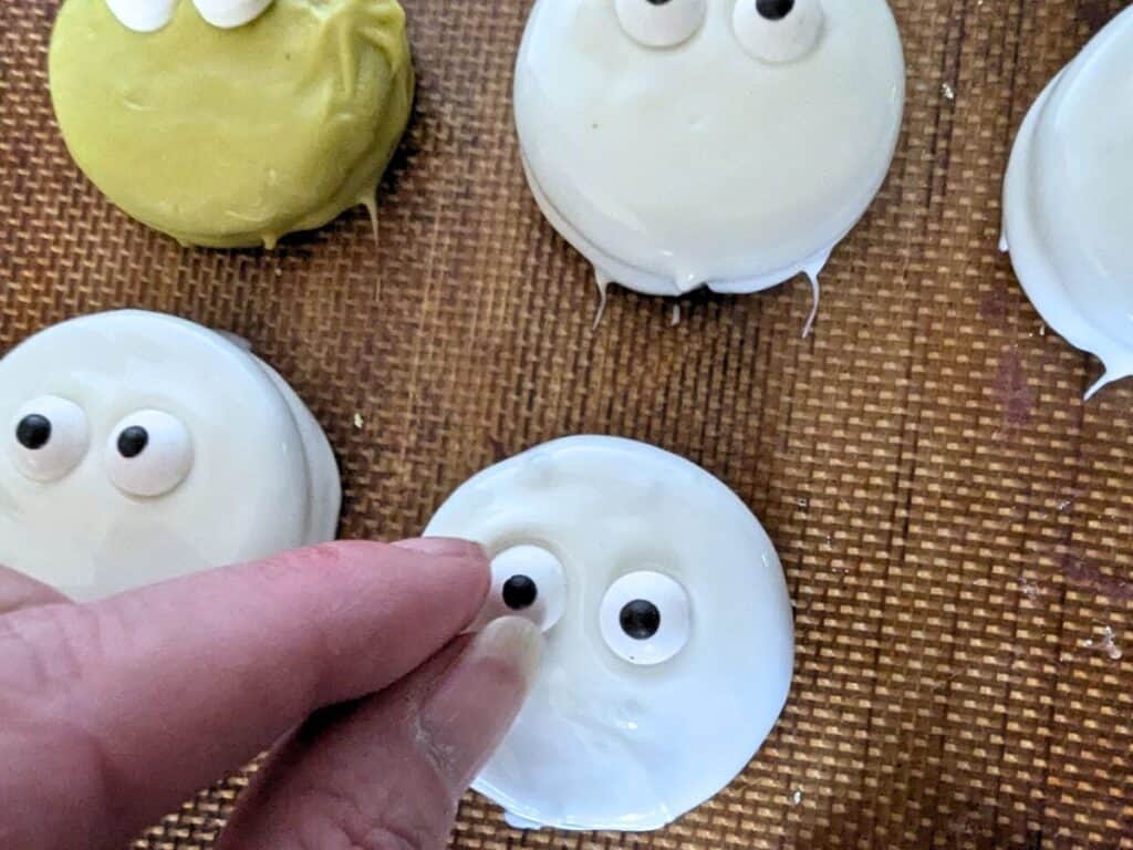 Image shows a Hand adding candy eyes to white chocolate dipped oreo.