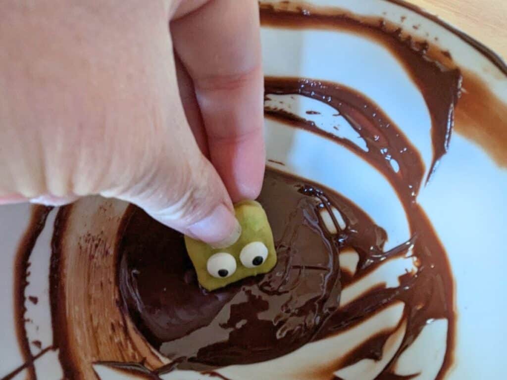 Image shows a Hand dipping a green pretzel into chocolate.
