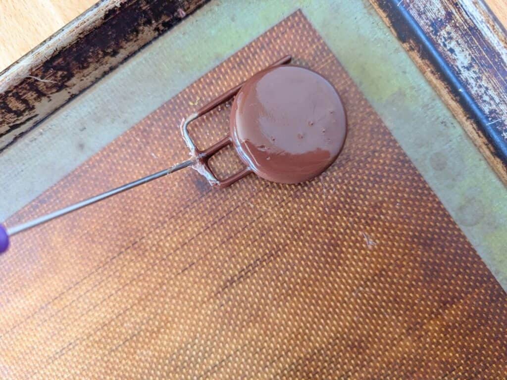 Image shows a chocolate dipping tool Sliding chocolate dipped cookie onto silpat.