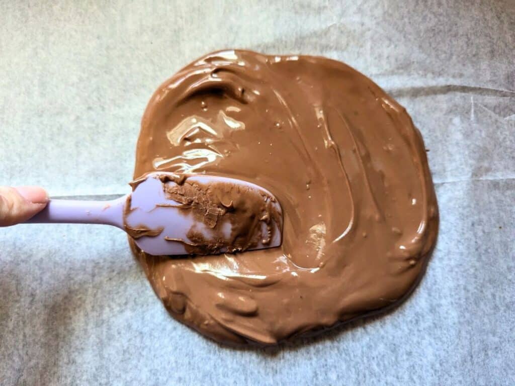 Image shows Spreading melted chocolate on parchment paper with a purple spatula.