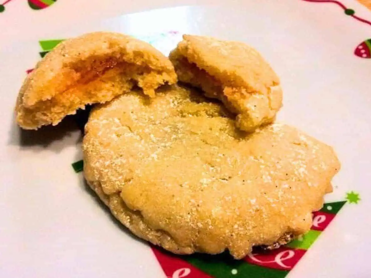 Image shows a sugar-dusted cookie with a bite revealing an orange filling, placed on a festive plate with holiday decorations.