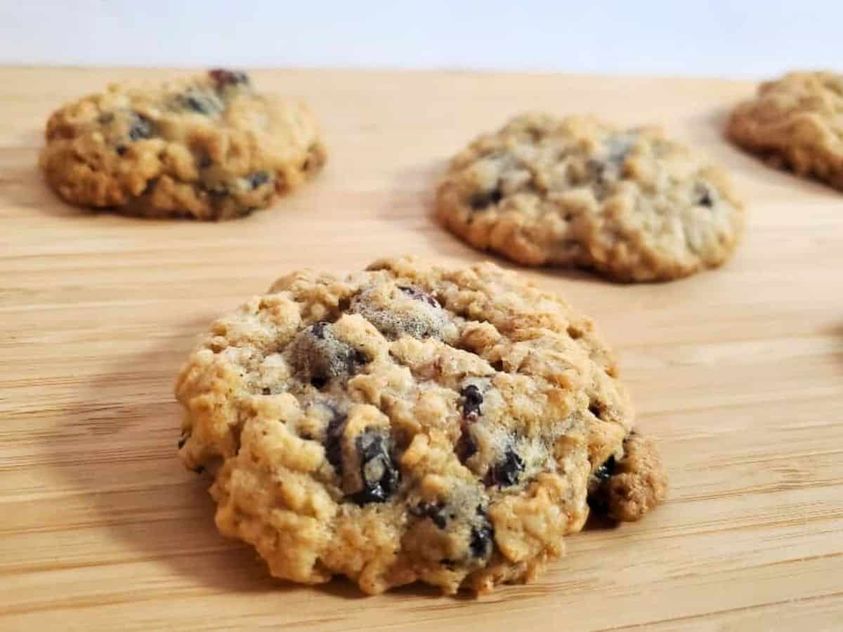Image shows hand holding an oatmeal raisin cookie with more cookies in the background on a wooden surface.