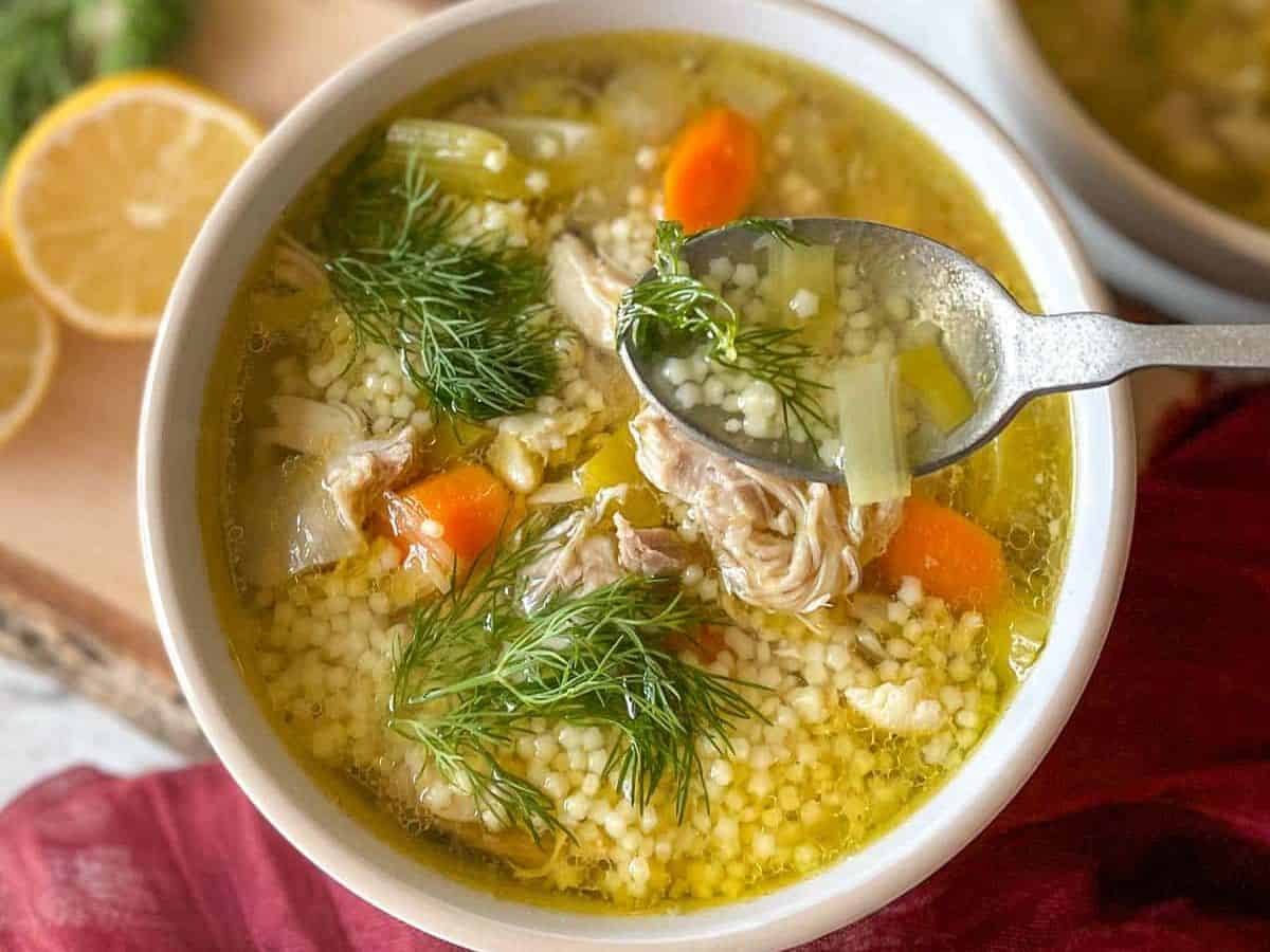 A bowl of chicken pastina soup sits on a rustic wooden cutting board with lemons and dill in the background.