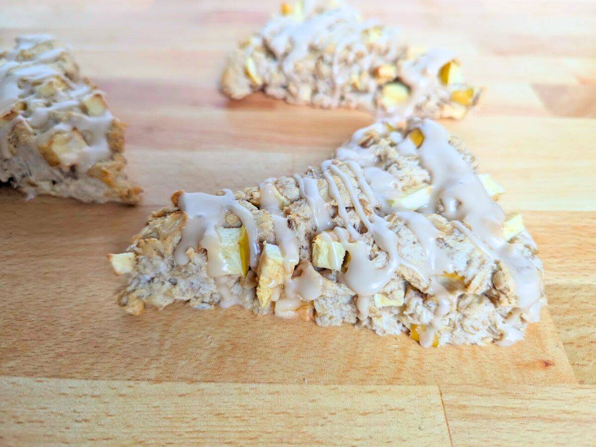 A triangular apple scone with visible oat and fruit pieces, topped with a light drizzle of white icing, on a wooden surface.