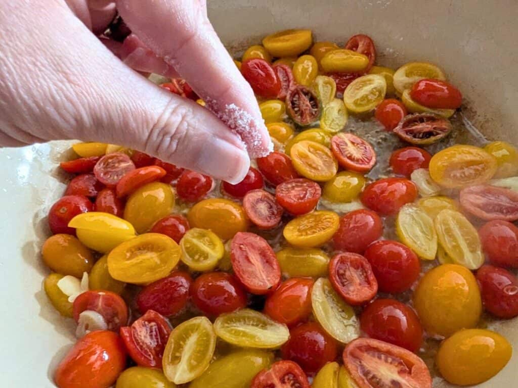 Image shows A hand adding salt to tomatoes simmering in a pan on the stove.