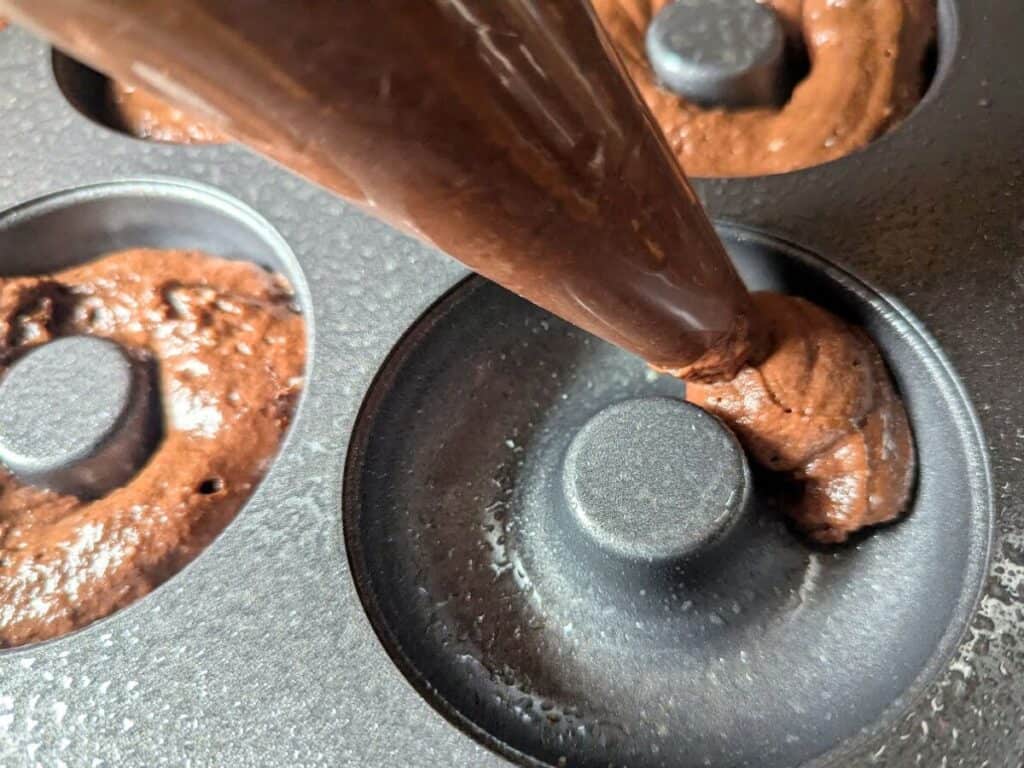 Image shows Chocolate donut batter being piped into a donut pan using a piping bag.