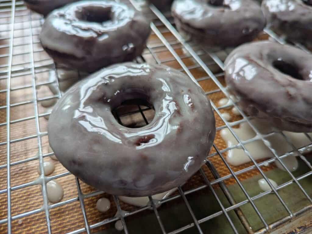 Image shows Glazed chocolate donuts on a cooling wire rack.