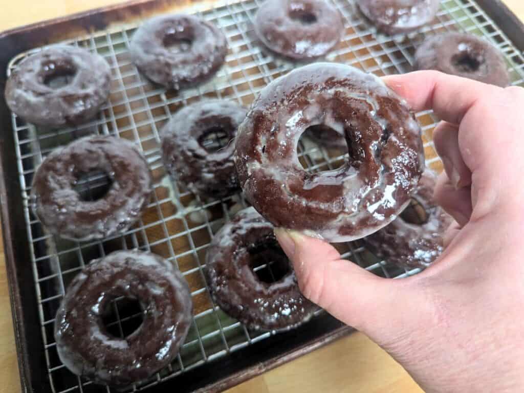 Image shows a Hand holding a glazed chocolate donut with more in the background.