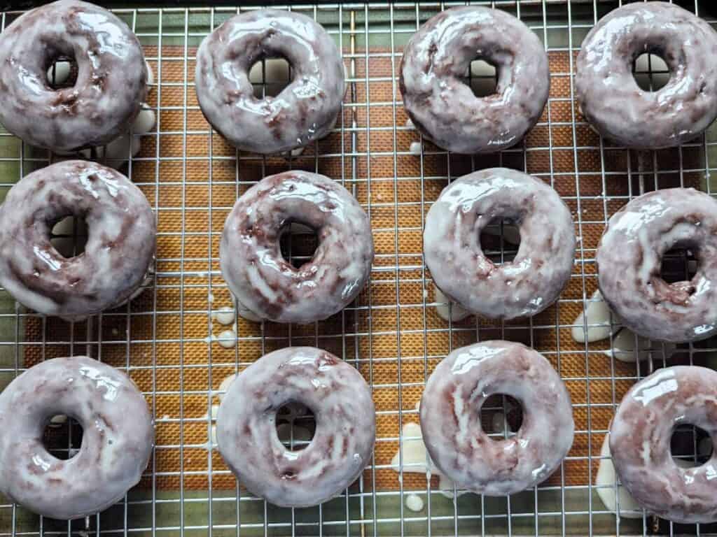 Image shows an Overhead view of glazed chocolate donuts cooling on a wire rack.