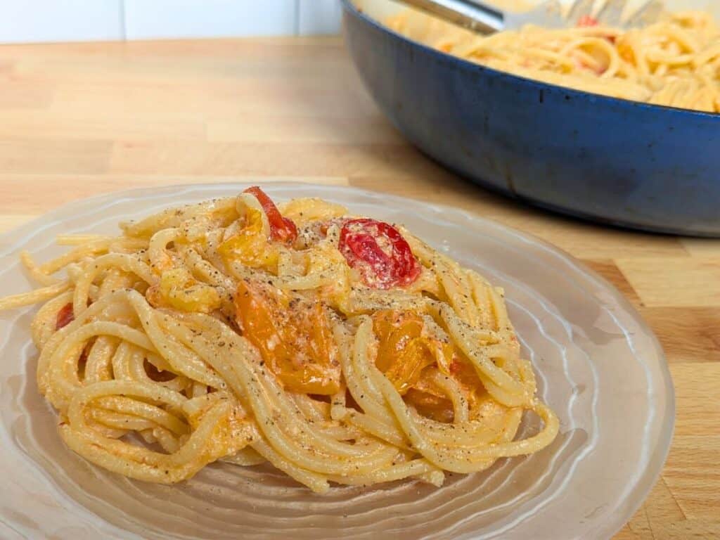 Image shows Plated pasta with tomatoes and ricotta ready to eat, with a pan filled with extra pasta behind it.