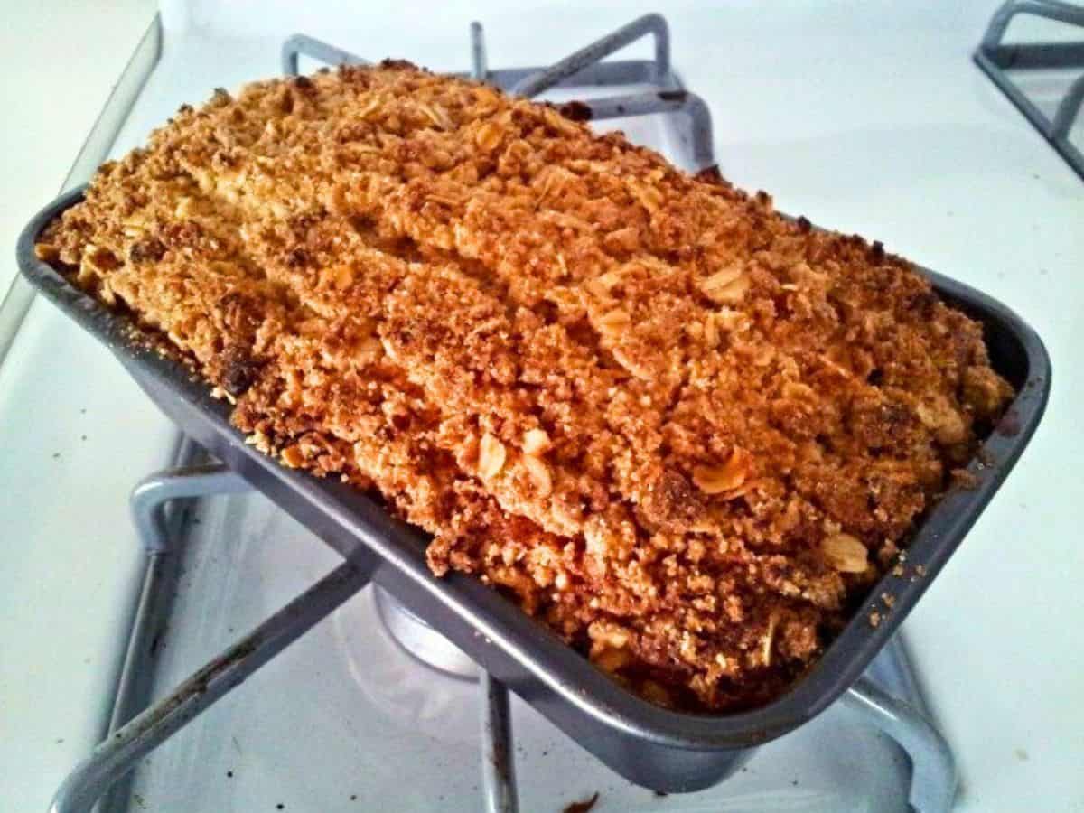 Image shows a loaf of baked bread with a golden brown, crumbly oat topping sits in a metal pan on a stove.
