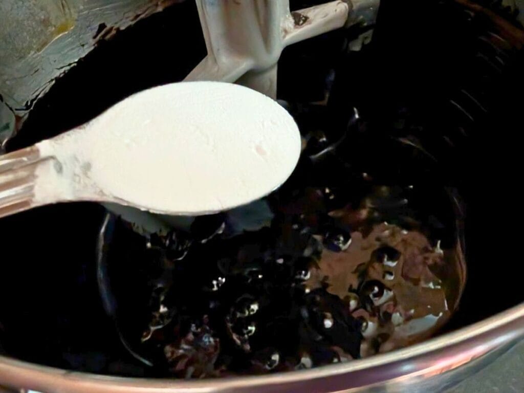 A white spoon holding baking soda hovers over a pot of boiling dark syrup, with bubbles forming on the surface.