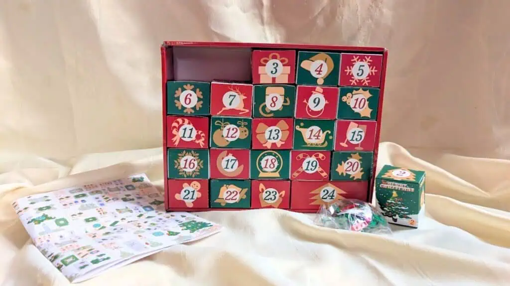 A festive Advent calendar box with numbered drawers, a sheet of stickers, a small decorated box, and a plastic bag with candies displayed in front—perfect for fans of Advent calendars.