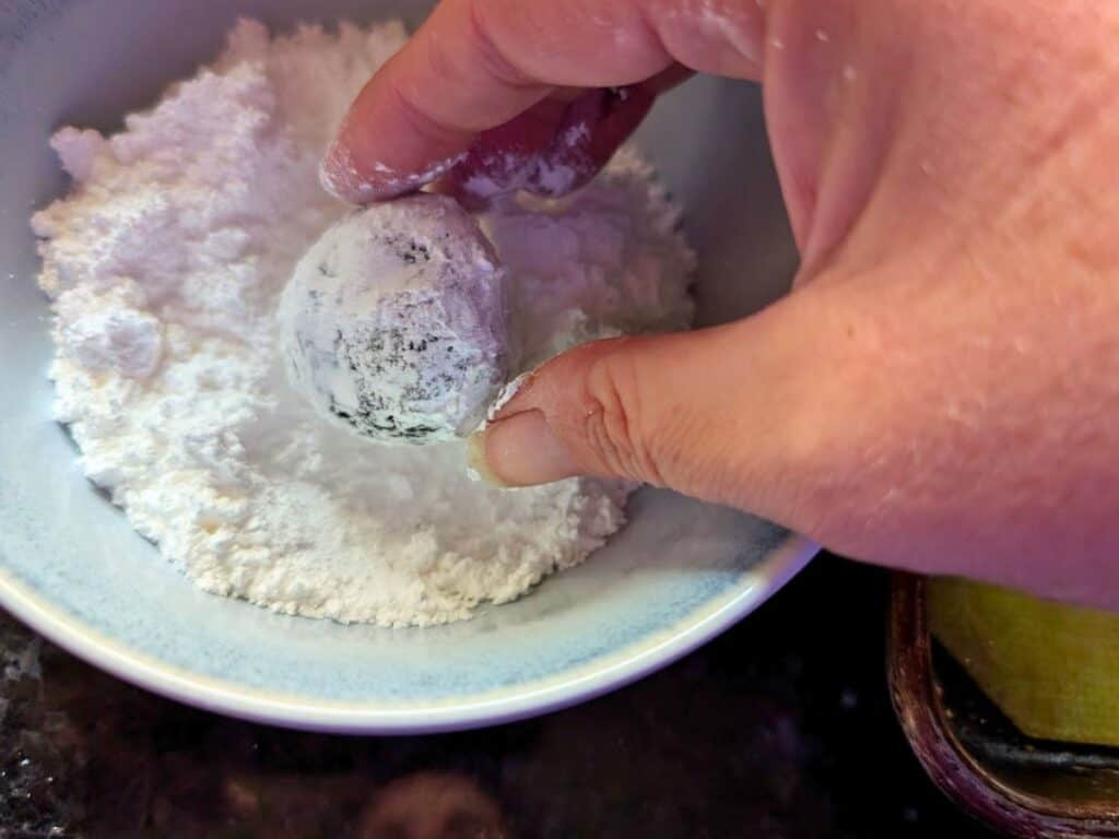 A hand coats a round chocolate truffle in powdered sugar inside a white bowl.