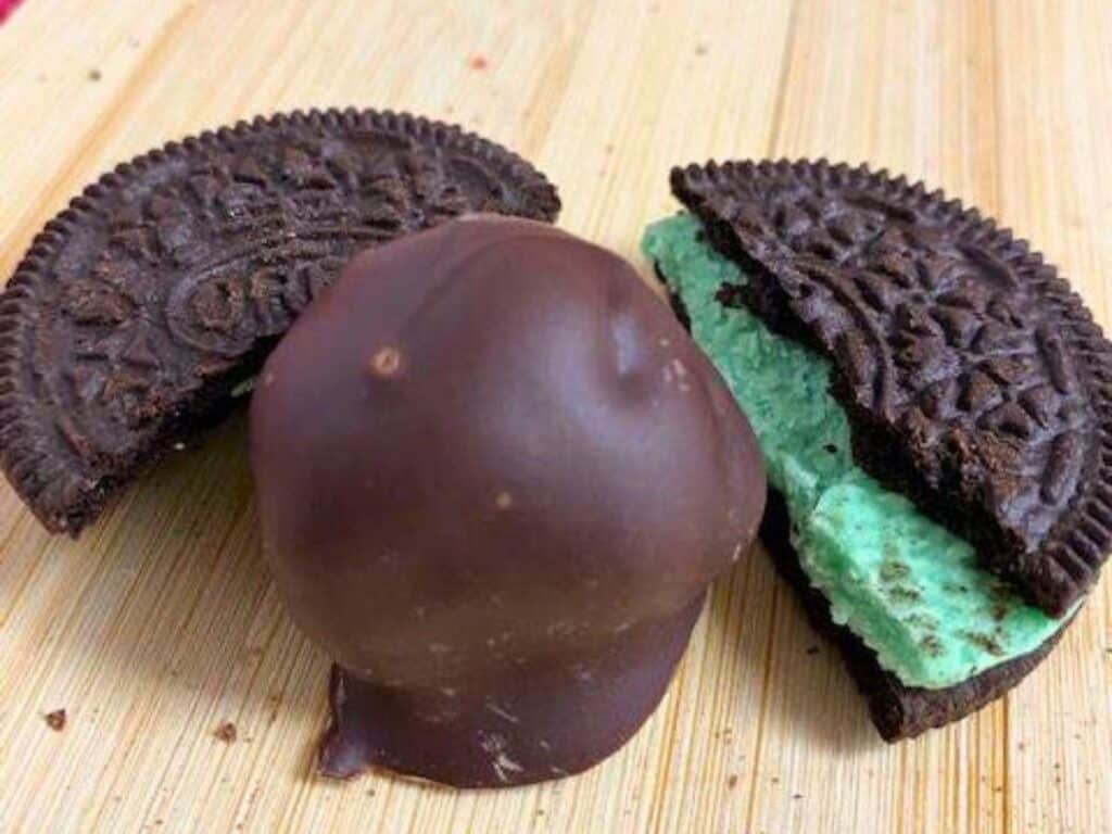 A chocolate-covered ball sits next to a mint cream-filled sandwich cookie split in half on a wooden surface.