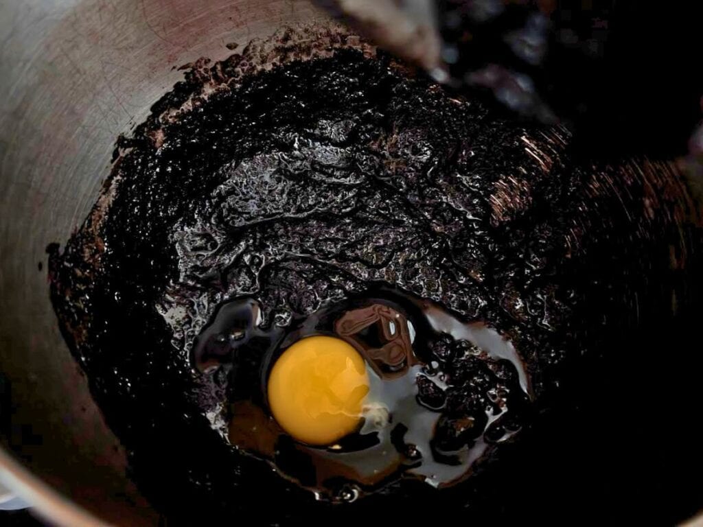 A raw egg sits on top of a mixture of dark, thick batter in a metal mixing bowl.