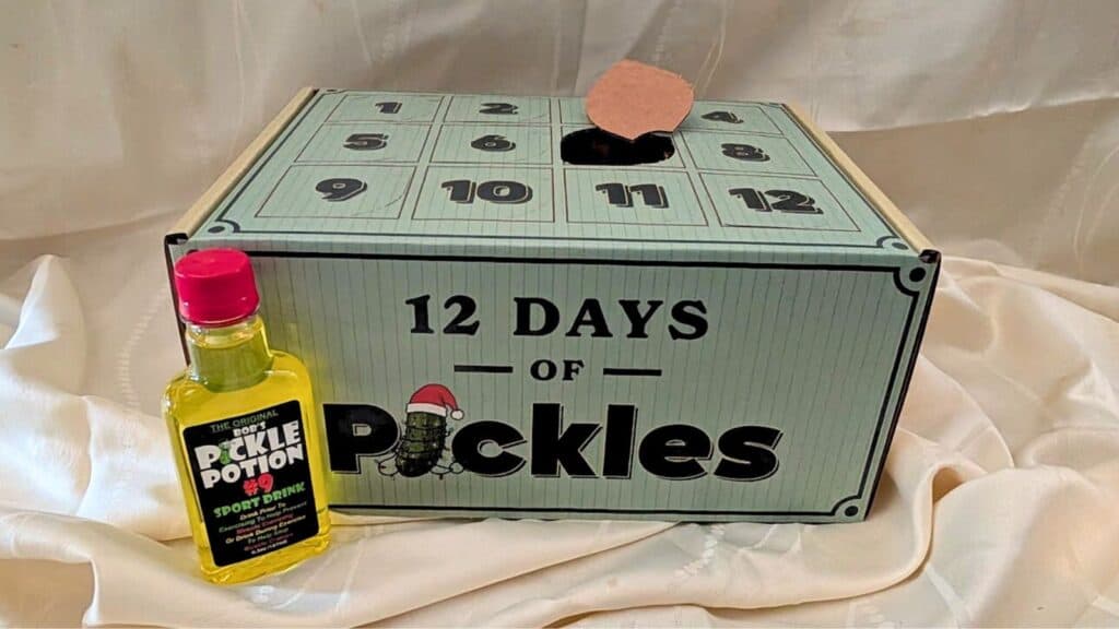 A box labeled “12 Days of Pickles” resembles quirky advent calendars, featuring numbered doors and a small bottle of pickle-flavored drink beside it, all set against a white fabric background.
